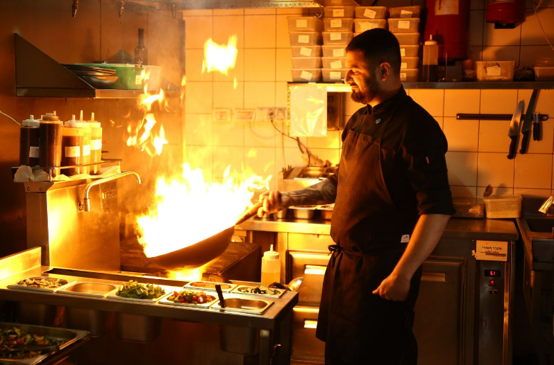 טבח בפס חם שמחזיק מחבת בלהבות במסעדת הביסטרו - מסעדה בשרית כשרה